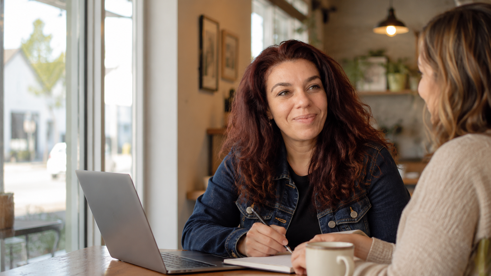 Two women talking over a laptop and a notebook at a cafe table